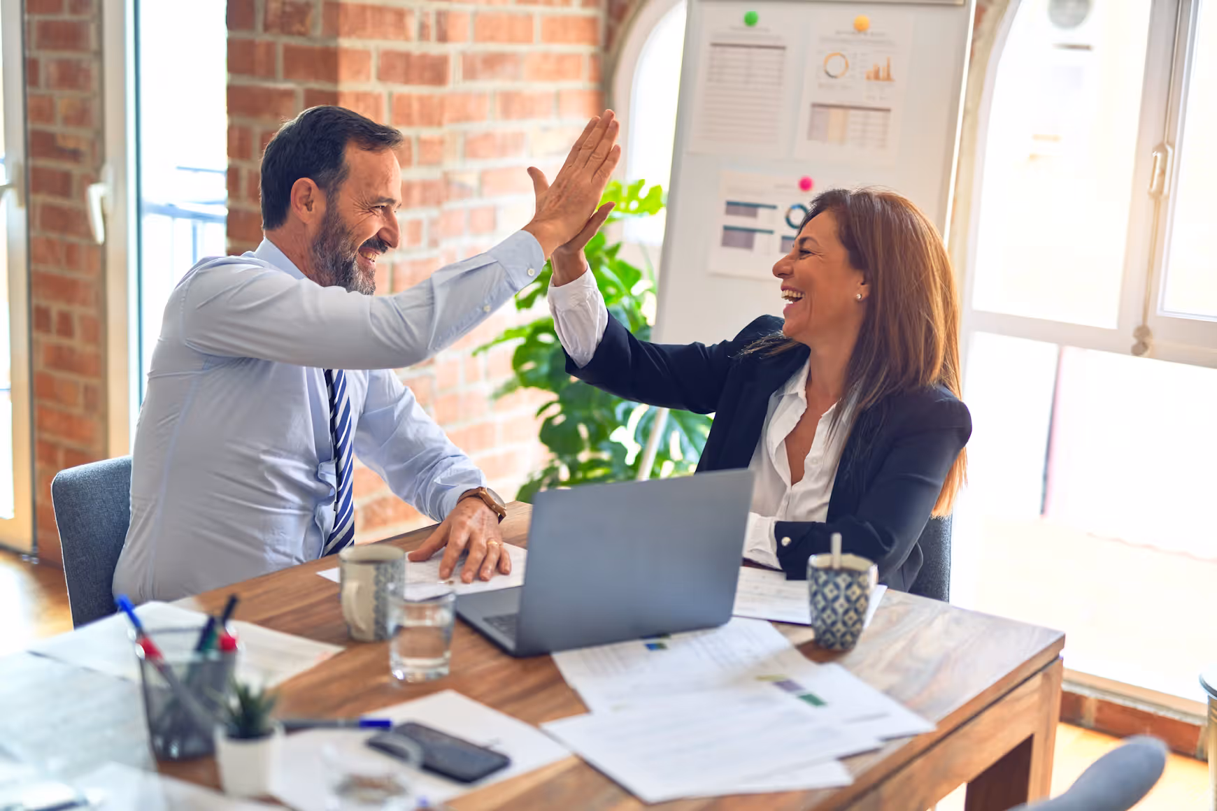 Two people high five while doing paperwork at a desk with a laptop.